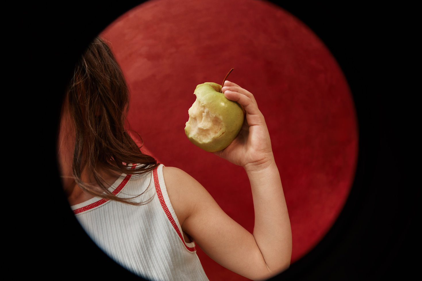 Child holding a green apple, viewed from behind, wearing a cream tank top with red trim against a red circular backdrop.