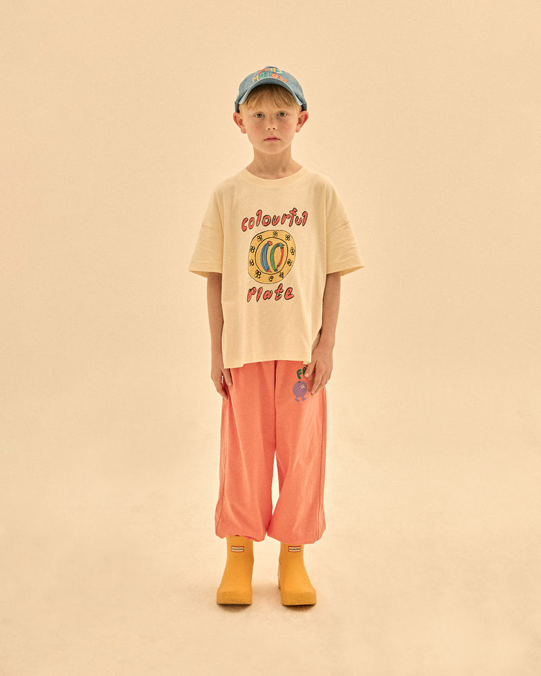 Child wearing a cream T-shirt with coral pants and a cap, standing against a neutral background