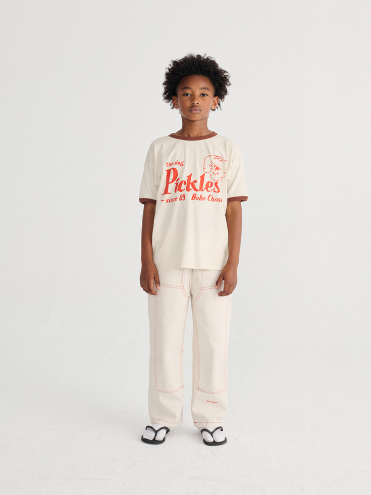 Child wearing cream pants with a white “Pickles” graphic T-shirt.