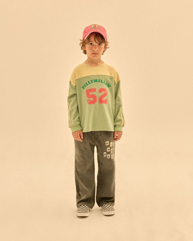 Child wearing dark denim pants with a green long-sleeve shirt and a cap, standing against a neutral background