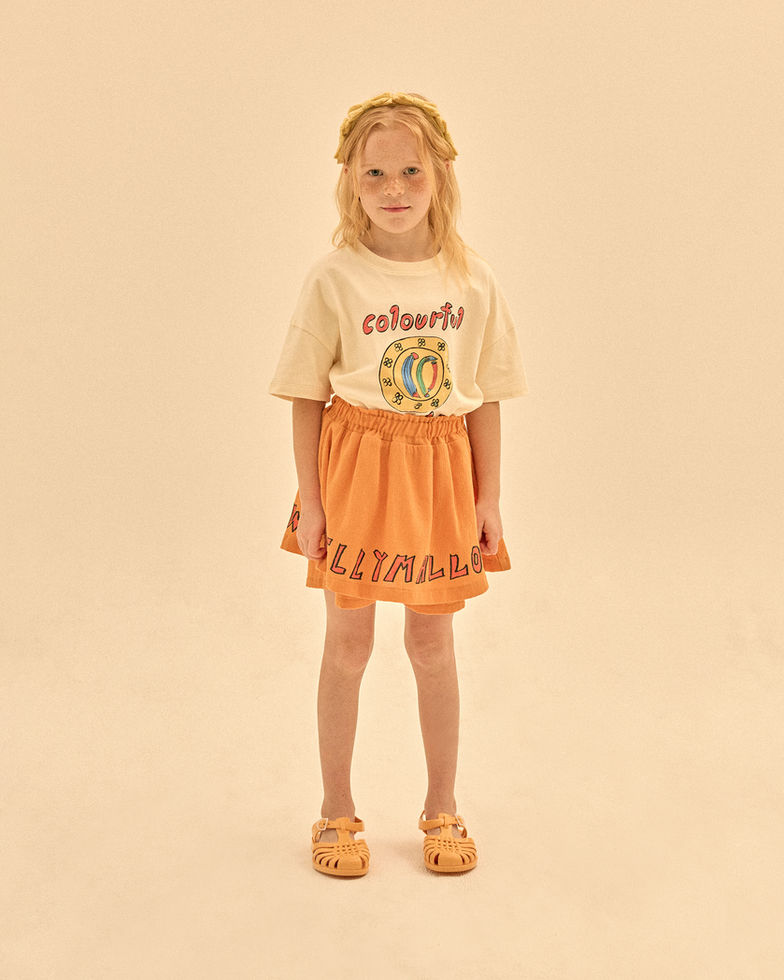Child wearing a cream T-shirt with an orange skirt, standing against a neutral background