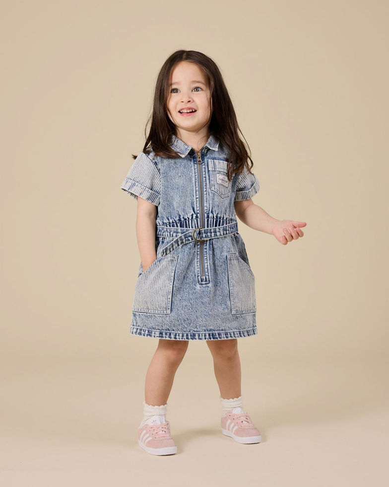 Young girl standing and smiling, wearing a short-sleeve denim dress against a beige background.