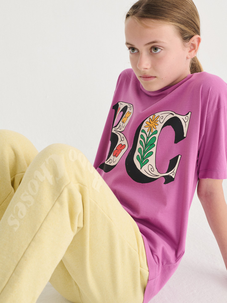 Close-up of a child wearing a pink “BC” T-shirt while seated, showing part of the graphic and yellow pants.