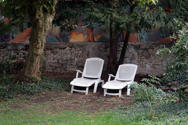 Félix Rouxel. Grandit la forêt. Acrylique. Juillet 2019. Un jardin arboré. Un meuret de pierre surmonté d'une fresque d'arbres. La fresqe est rouge jaune et bleu ciel. Elle représente des troncs, épais et tordus devant des feuillages denses et des épis floraux dressés.