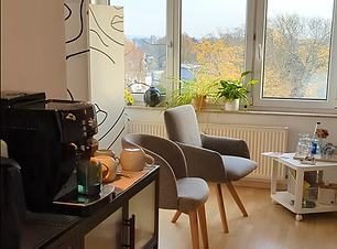 Ein Auschnitt aus dem Praxisraum, man sieht vorne links eine Kaffeemaschine auf einem Sideboard, dahinter stehen zwei graue, bequem aussehende Stühle vor einem Fenster, daneben ein kleiner Tisch mit Getränken.