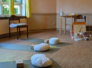 Auschnitt des Raumes, in dem die Geburtsvorbereitungskurse stattfinden, man sieht drei Matten mit jeweils einem Kissen und einem Klotz, an der Wand steht in Stuhl und rechts im Bild steht eininges an Anschaungsmaterial