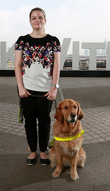 A lady stands on concrete with her guide dog sitting beside her.