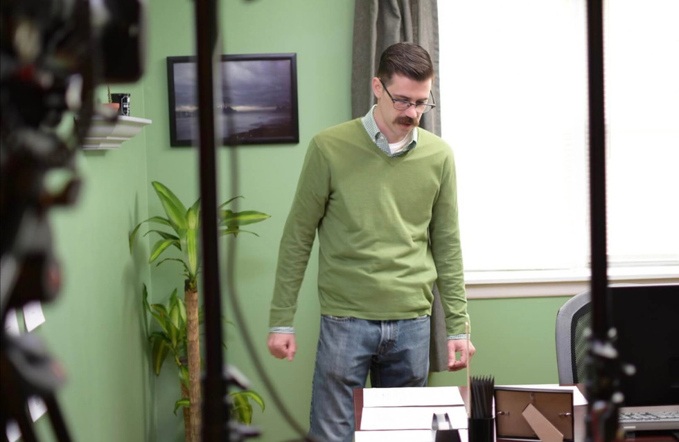 a man wearing glasses and a green sweater stands in front of a window
