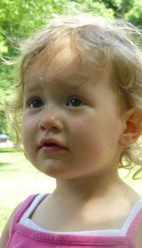 A close-up portrait captures a young child with curly blonde hair looking off to the side with a wistful expression. The child, dressed in a pink top, is situated in a sunlit outdoor setting with blurred green trees and grass in the background.