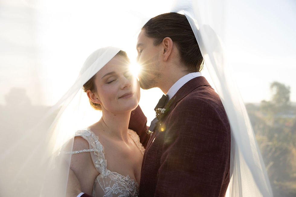 Groom kissing bride on the cheek under her veil, her eyes are closed, the sun is flaring between their faces.