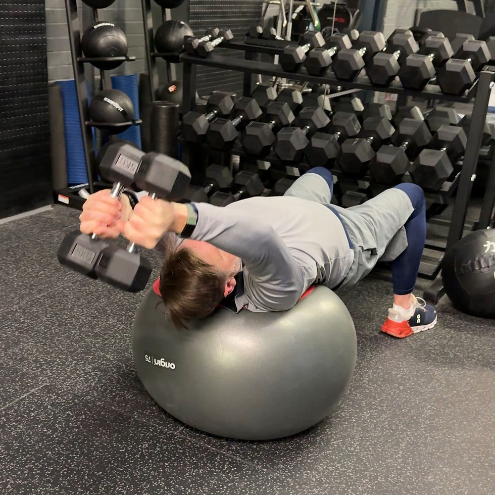 Man in gray workout gear lies on exercise ball lifting dumbbells in a gym. Dumbbells and medicine balls on racks in the background.