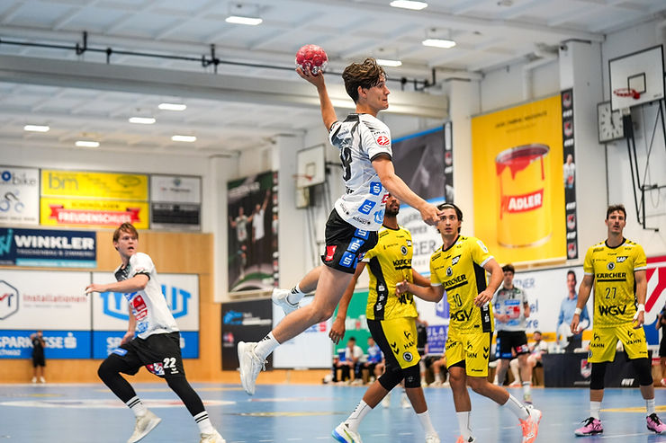 Handballspieler im Sprung mit Ball, im Hintergrund Gegenspieler in gelben Trikots