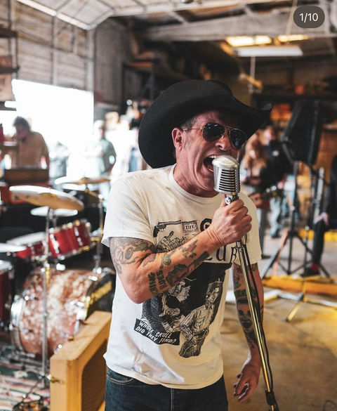 A tattooed musician in a cowboy hat singing into a vintage microphone.