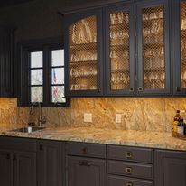 Luxury custom home bar and beverage station by Birdy Design Studio featuring dark bespoke cabinetry, illuminated glass-front cabinets with decorative brass mesh, warm natural stone countertops and backsplash, integrated bar sink, and curated glassware display for an elevated luxury interior in Tulsa, Oklahoma.