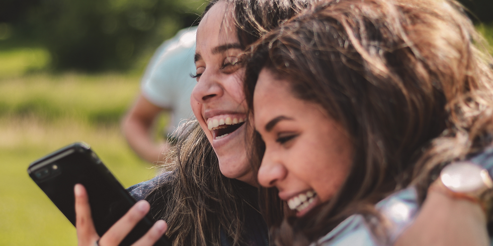 Two laughing women share a phone, smiling together in an outdoor setting.