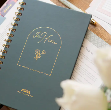 A dark teal spiral-bound 'In Him' prayer journal with gold foil lettering, styled with flowers.