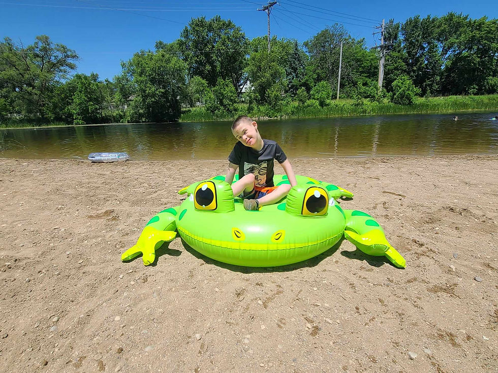 Jasper sitting on a large inflatable frog at the beach, enjoying a sunny day by the water with a big smile on his face.
