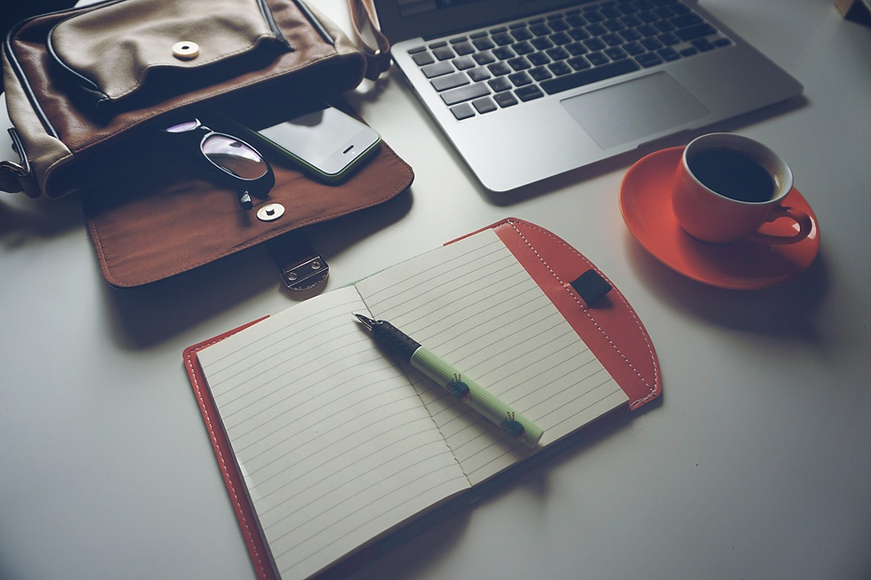 Open notebook, pen, laptop, bag, phone, glasses, and coffee cup on a desk. Warm lighting creates a cozy, productive atmosphere.