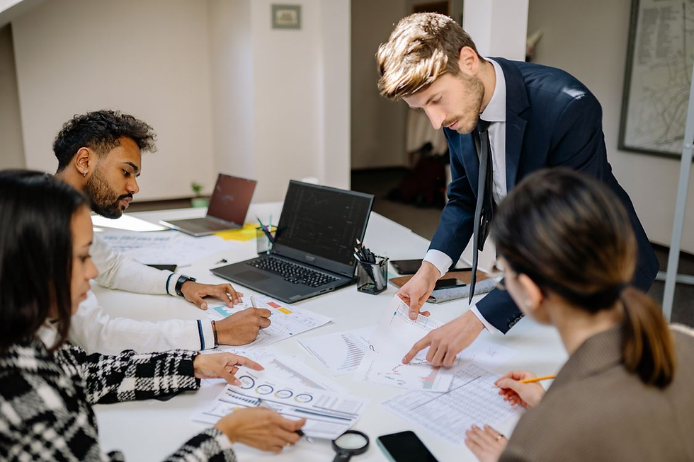 Business team reviewing charts at a white table, with a man in a navy suit pointing at reports in a bright office.