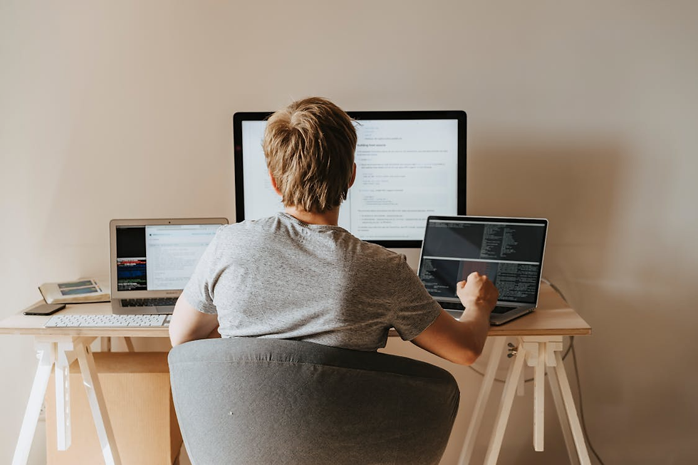 Person in a gray shirt working on three screens displaying code at a wooden desk. Bright room with a minimalist setup.