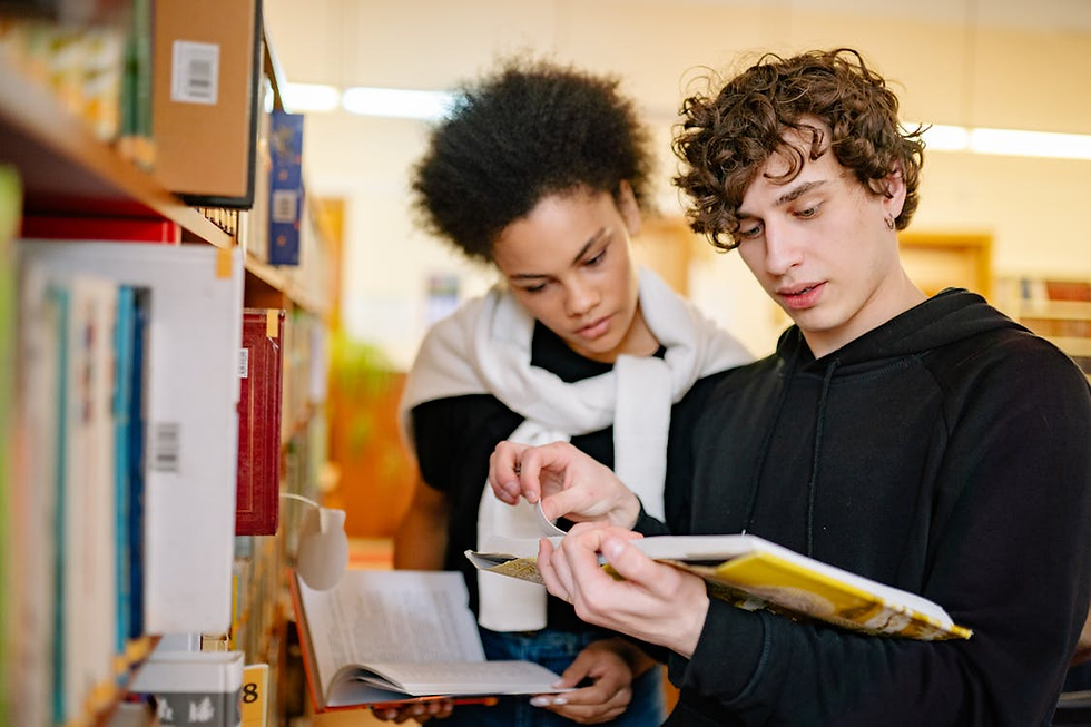 Two students in a library browse books together, one pointing at a page while both read in a focused, quiet setting.