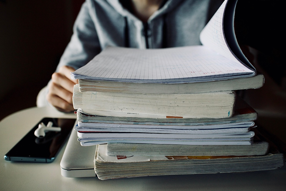 Stack of books and a notebook on a white table. A person in a gray hoodie turns a notebook page. A smartphone with earbuds is nearby.
