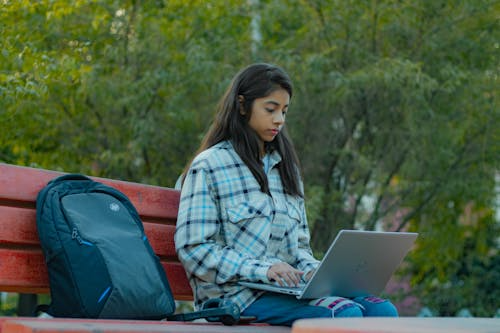 Young woman in plaid shirt using laptop on a red bench with black backpack nearby; set in a green, leafy park.