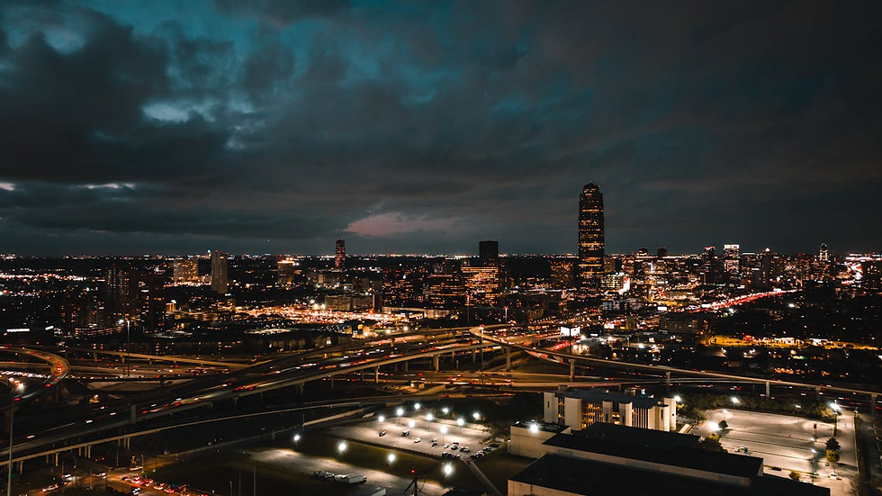 City skyline at dusk with illuminated buildings, a towering skyscraper, and busy highways. Dark clouds create a dramatic, moody atmosphere.