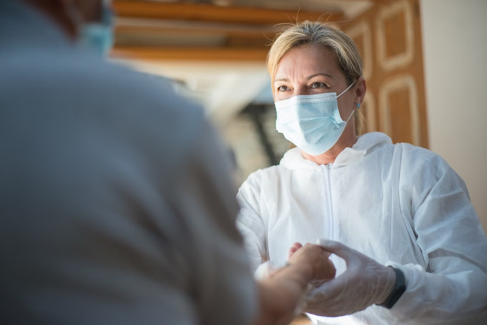 Masked woman in white protective suit shakes hands with a blurred person indoors, looking reassuring.