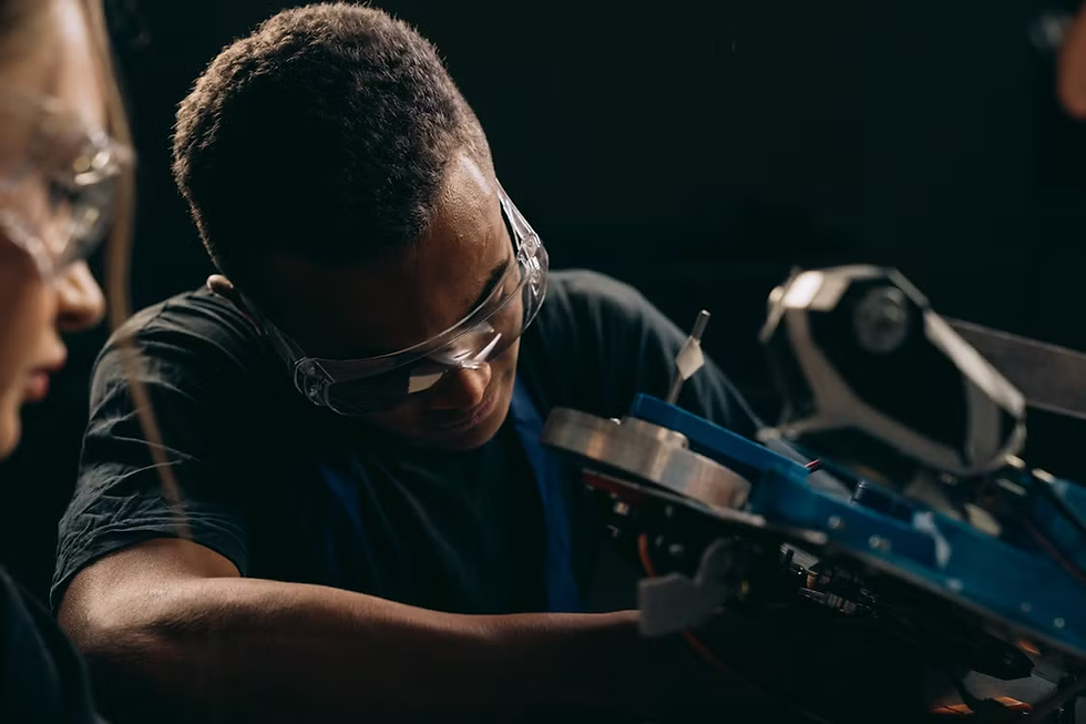 Teen in safety goggles works on a machine in a dark workshop, with another person watching nearby.