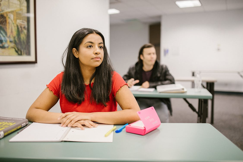Woman in red shirt attentively listens in classroom, with a man in background. Green desks, books, and a pink organizer are visible.
