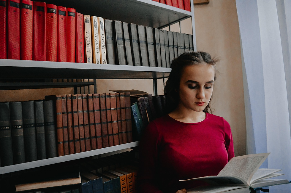 Woman in red sweater reading a book in a library. Background features shelves filled with red, brown, and gray books. Calm setting.