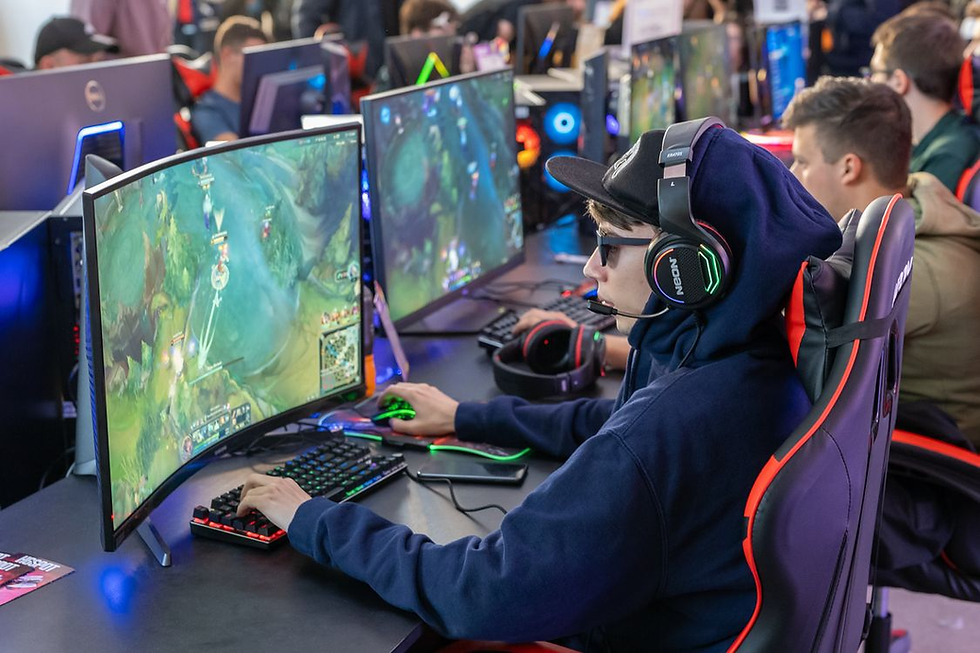 Teen gamer in hoodie and headset plays on a curved monitor at an esports event, with rows of glowing PCs behind him.