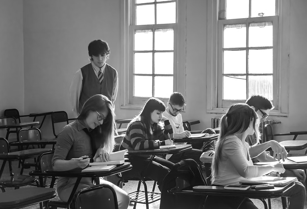 Black-and-white classroom with students writing at desks while a young man stands behind them near bright windows.