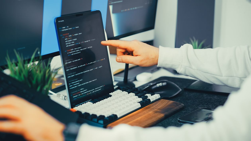 Person points at a tablet showing code beside a keyboard and monitor in a modern desk setup, focused and technical