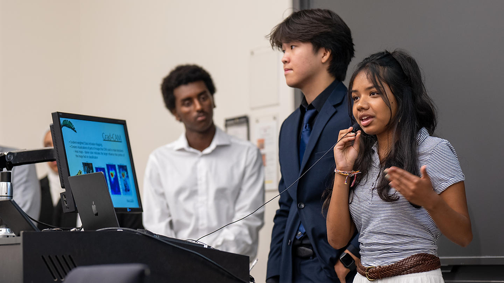 Three people present in a classroom. A woman speaks into a mic. A screen displays "CAD-CAM" with images. The mood is engaging.