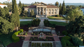 Grand building with columns, surrounded by trees and garden. "REDLANDS" spelled in greenery. A few people walk nearby. Calm, sunny day.