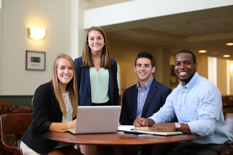 Four people in business attire smiling at a table with a laptop, papers, and pens. Bright modern office setting, warm lighting.