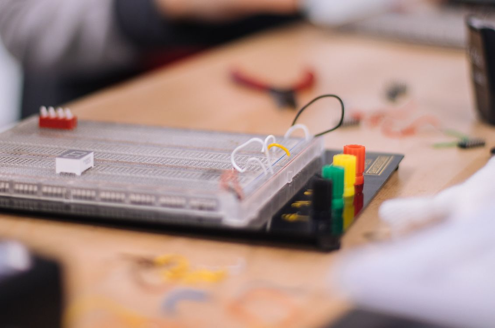 Breadboard electronics project on a worktable, with colorful connectors and wires, blurred tools and hands in the background.