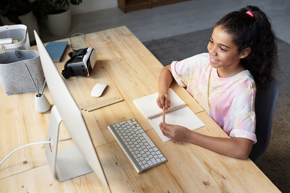 Girl in tie-dye shirt smiling at a computer, writing in a notebook at a wooden desk with VR headset, keyboard, and books. Bright, cheerful setting.