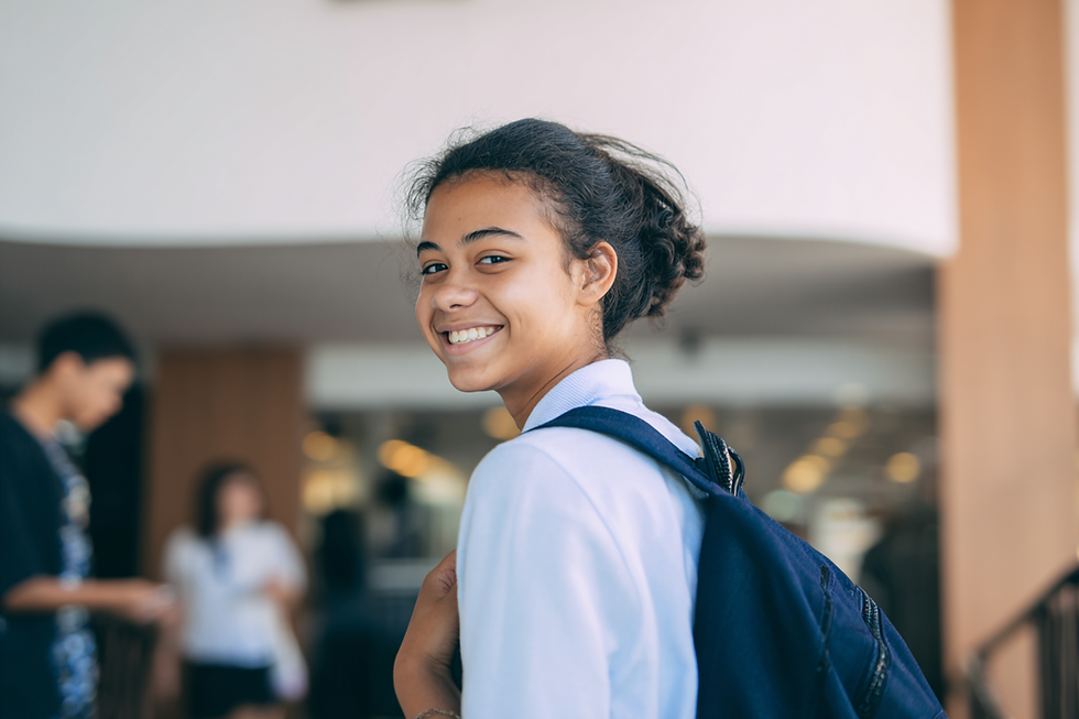 Smiling person in a light blue shirt with a navy backpack, standing indoors. Blurred background with people and soft lighting.