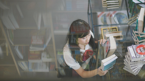 Young woman browsing books in a library, surrounded by stacked books. Reflections and soft lighting create a calm and focused mood.