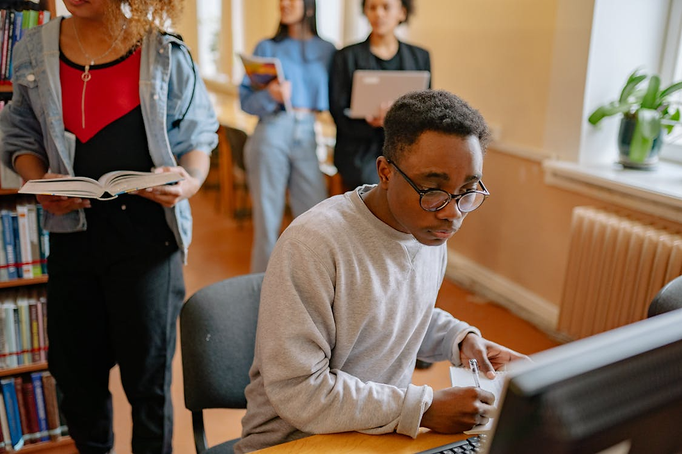 Students study in a library as a young man writes at a computer, while others stand reading books in the warm classroom.
