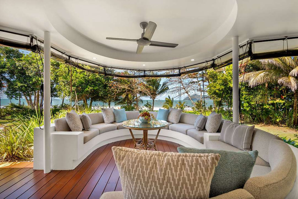 Open-air tropical lounge with curved white sofa and glass table, overlooking lush trees and the ocean. Reid road property