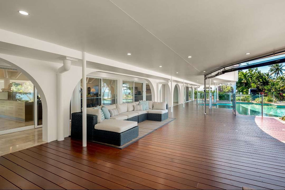 Spacious covered patio with white sectional on a wood deck beside a turquoise pool and tropical garden, bright and airy.