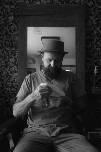 Ben the barber in Cumberland in a black and white photo sitting in his chair he cuts in wiping his glasses clean wearing a top hat