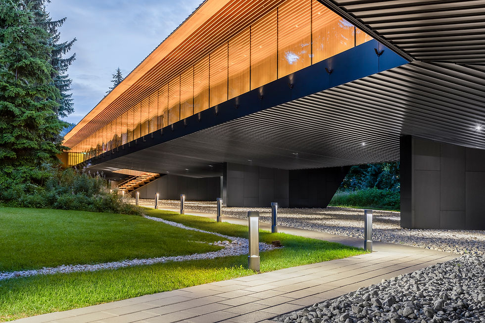 Modern building with a wooden facade, elevated on pillars over a lawn. Pathway lights illuminate the area. Surrounded by trees, with a serene mood.