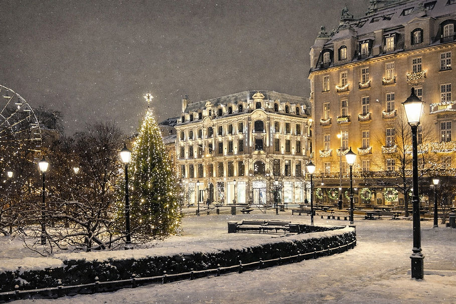 Akvarellillustrasjon av et snødekt torg i Oslo en stemningsfull vinterkveld, med opplyst juletre, historiske bygårder og varme gatelykter som speiler seg i snø og brostein. Myke snøfall, julelys og klassisk arkitektur skaper en rolig og nostalgisk stemning fra vinterbyen, med et hint av pariserhjul i bakgrunnen og mennesker som beveger seg gjennom det opplyste byrommet.