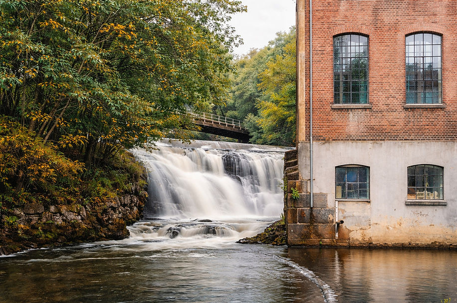 Fossefall og eldre industribygg langs Akerselva i Oslo, omgitt av grønt landskap og historiske fabrikkområder – et motiv som forteller historien om industri, energi og byutvikling i Norge.