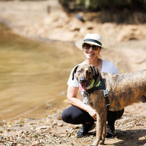 mulher de cnapéu e óculos escuros, sorrindo,  com cachorro na beira de um lago